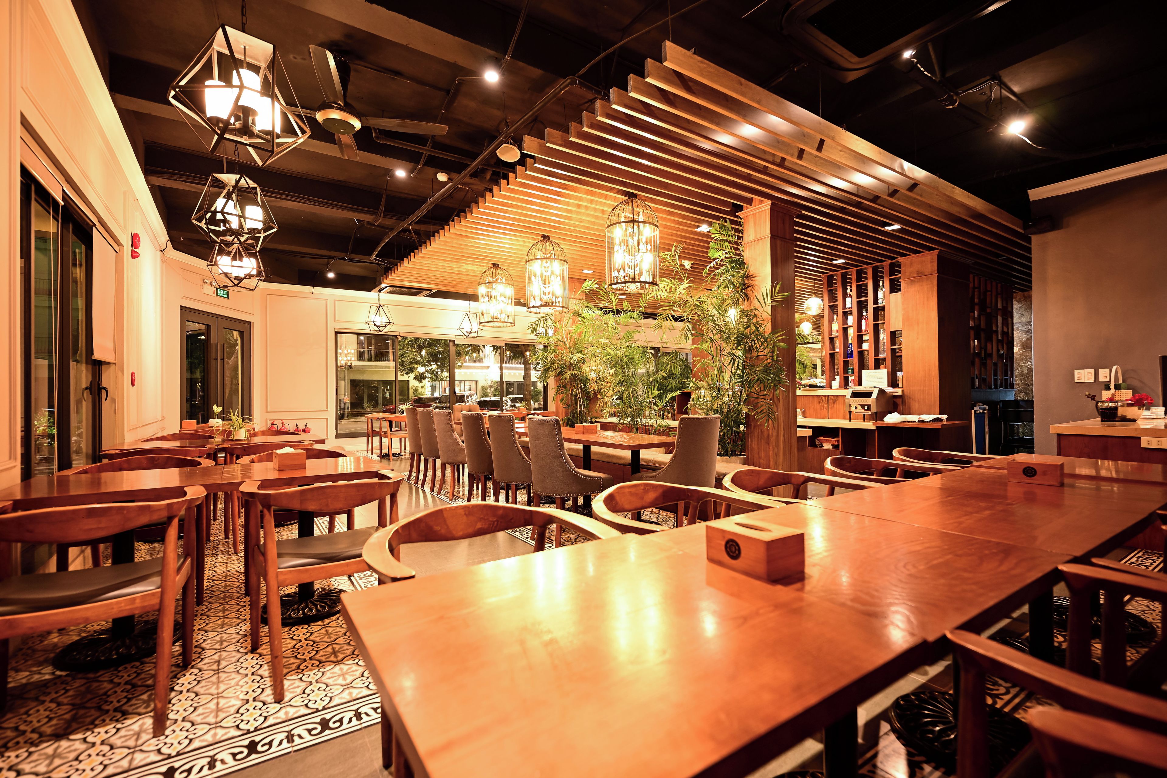 A restaurant interior with wooden tables and chairs, hanging lights, and a central plant.