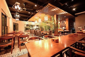 Interior de restaurante com mesas e cadeiras de madeira, luminárias pendentes e uma planta central.