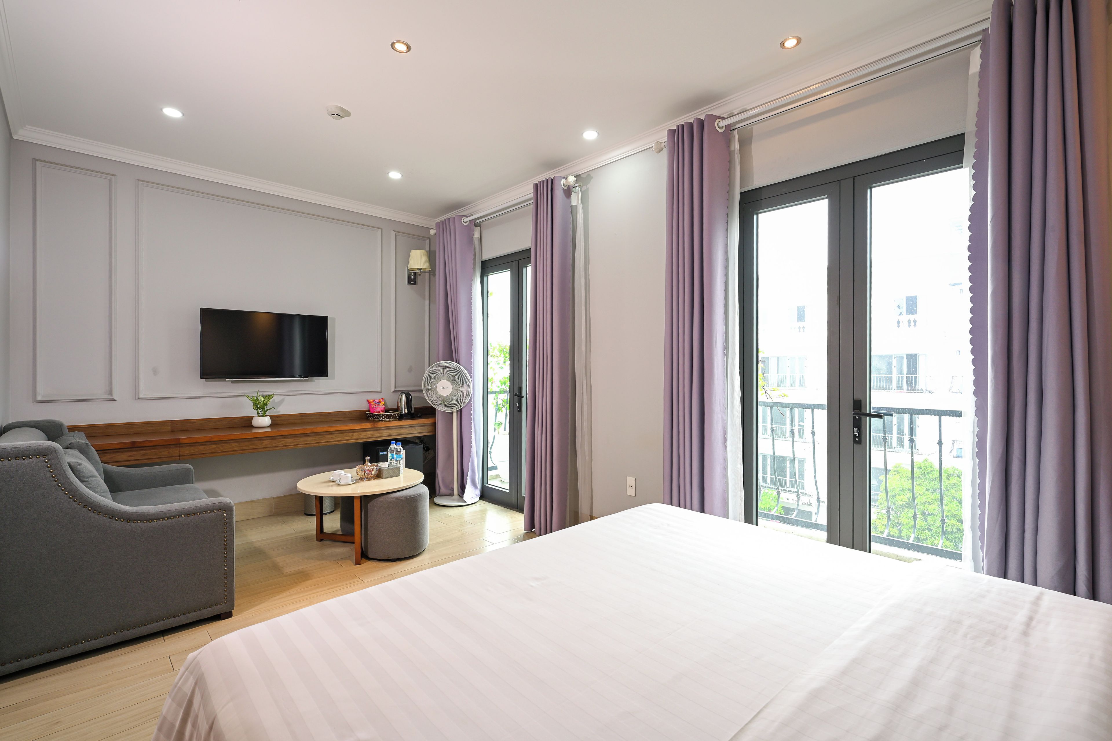 A hotel room with a bed, a seating area with a chair, a small table, a TV mounted on the wall, and a balcony with curtains.