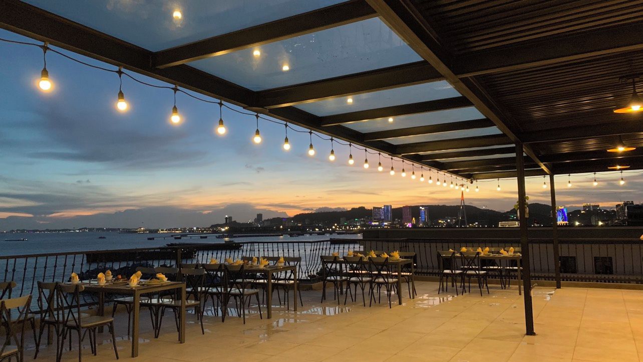 Un área de comedor al aire libre con mesas y sillas, luces colgantes y vistas al agua y al horizonte de la ciudad.