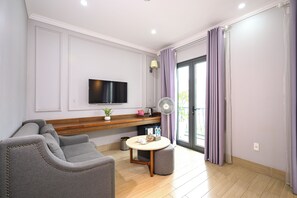 A modern living room with a grey sofa, a flat-screen TV mounted on the wall, a small table with a fan, and a sliding glass door with purple curtains. - The Confetti Hotel Ha Long (Hong Gai)