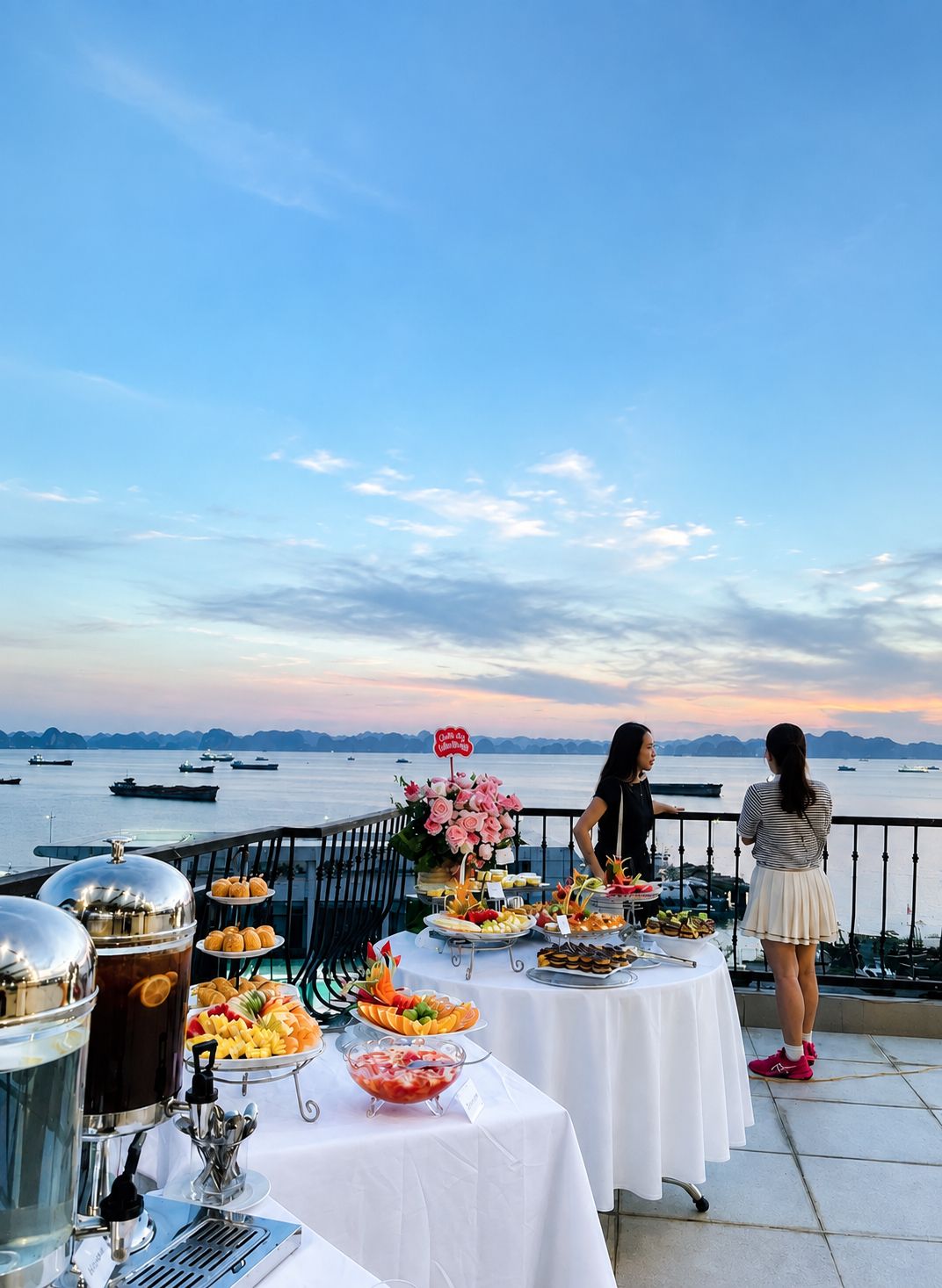 A buffet table with various food items and a coffee station, set against a backdrop of a body of water and distant mountains.