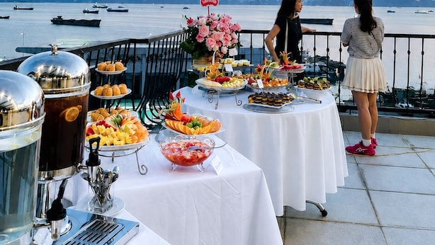 A buffet table with various food items and a coffee station, set against a backdrop of a body of water and distant mountains. - The Confetti Hotel Ha Long (Hong Gai)