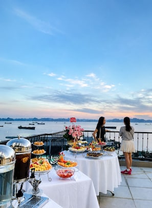 Mesa de buffet com diversos alimentos e um espaço para café, com vista para um corpo d'água e montanhas ao longe.