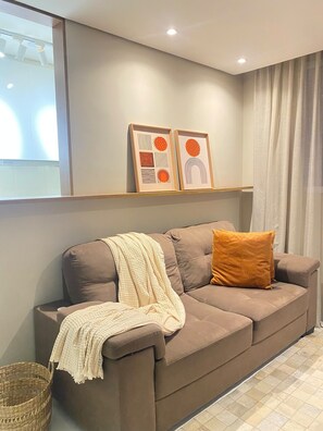 Una sala moderna con un sofá marrón, una manta blanca y una única almohada naranja. Hay dos obras de arte enmarcadas en la pared sobre el sofá.