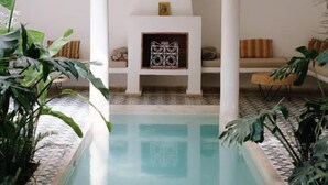 A pool area with a white tiled floor, a seating area with cushions, and a decorative wall with a framed picture. - Riad L'Atelier (Marrakech)