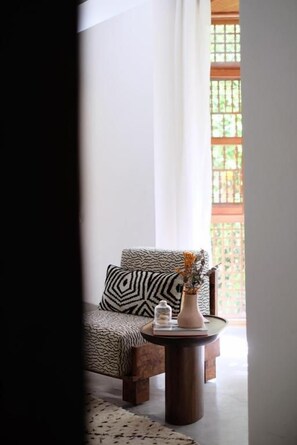 A room with a patterned sofa, a small round table with a vase and a book, and a view through a window with a lattice design. - Riad L'Atelier (Marrakech)