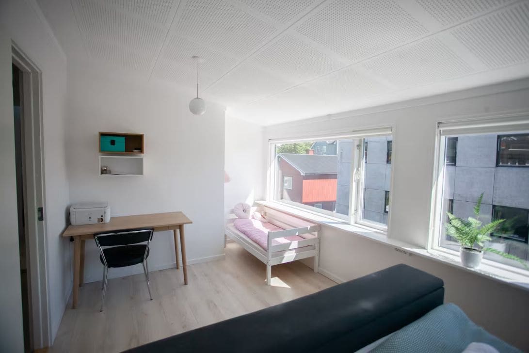 Ett rum med en säng, ett skrivbord, en stol och ett fönster med utsikt över byggnader.