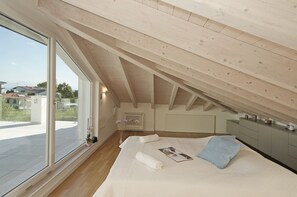 A bedroom with a sloped ceiling, a large window, a bed with pillows and a rolled towel, a bedside table, and a dresser. - Villetta I Giardini del Garda (Peschiera del Garda)
