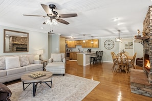 A spacious living room with a fireplace, a dining area with wooden chairs, a kitchen with wooden cabinets, and a ceiling fan.