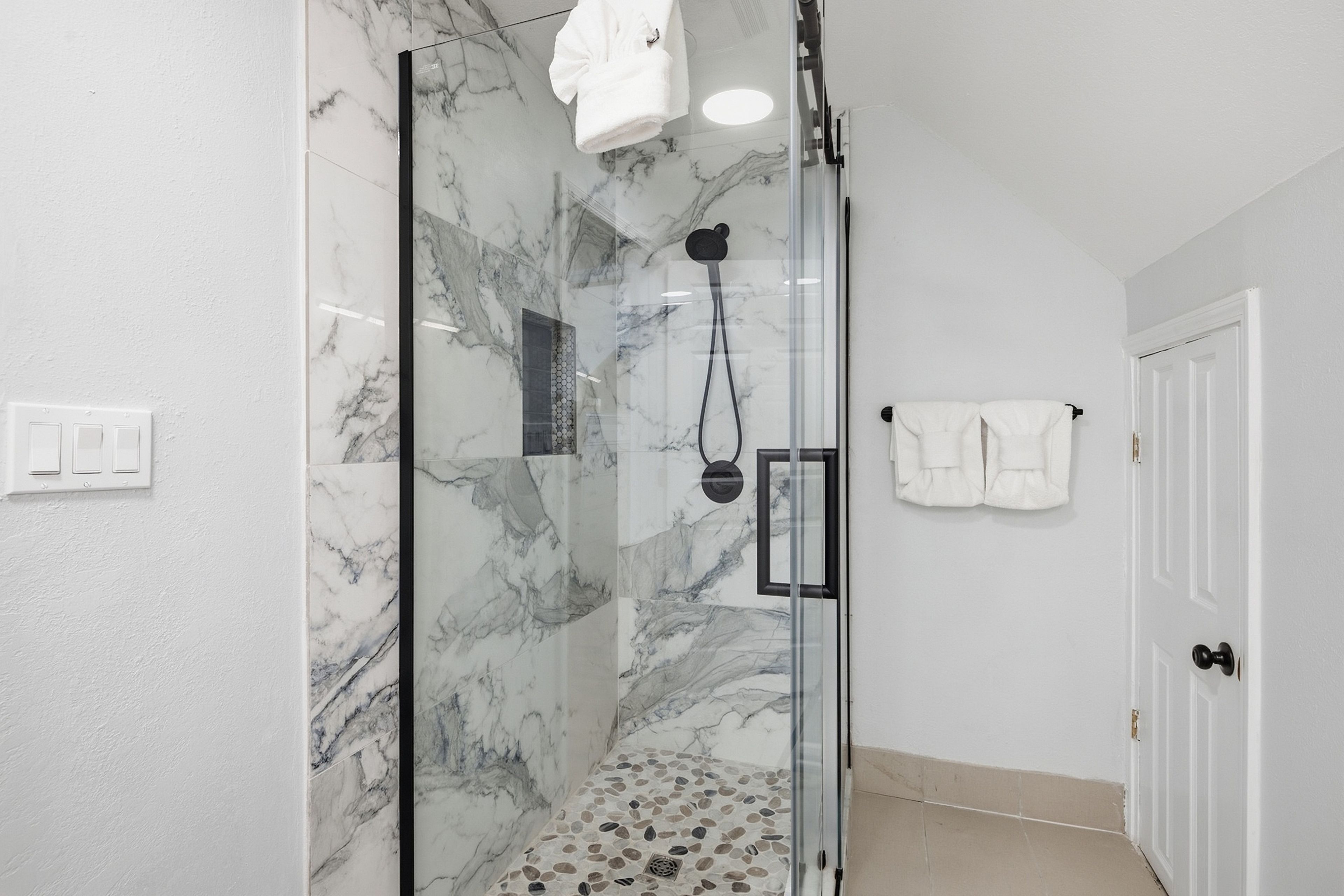 A glass-enclosed shower with marble walls, a white towel hanging on the wall, and a door with a towel rack.