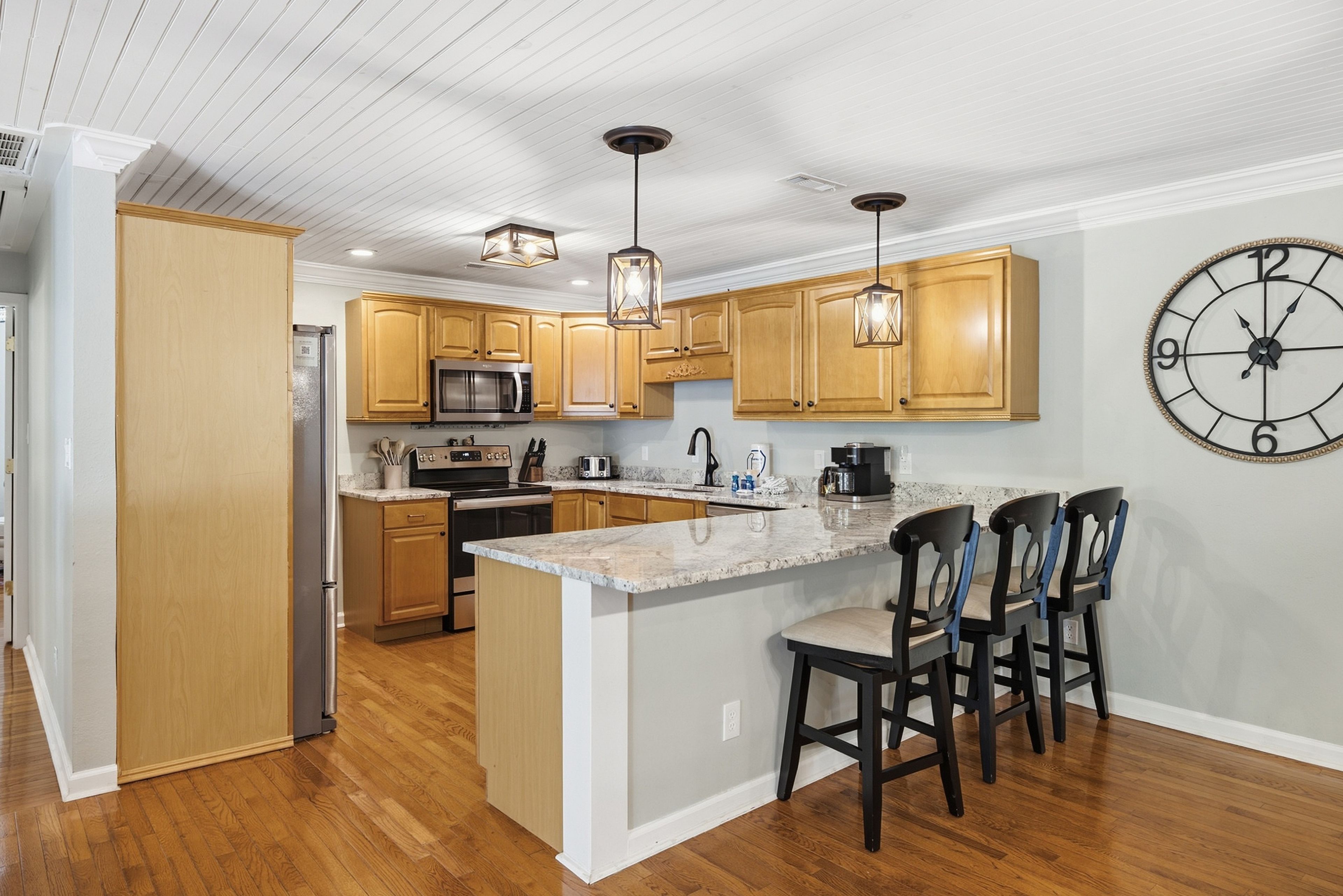A modern kitchen with wooden cabinets, a central island with bar stools, a built-in microwave, and a large wall clock.