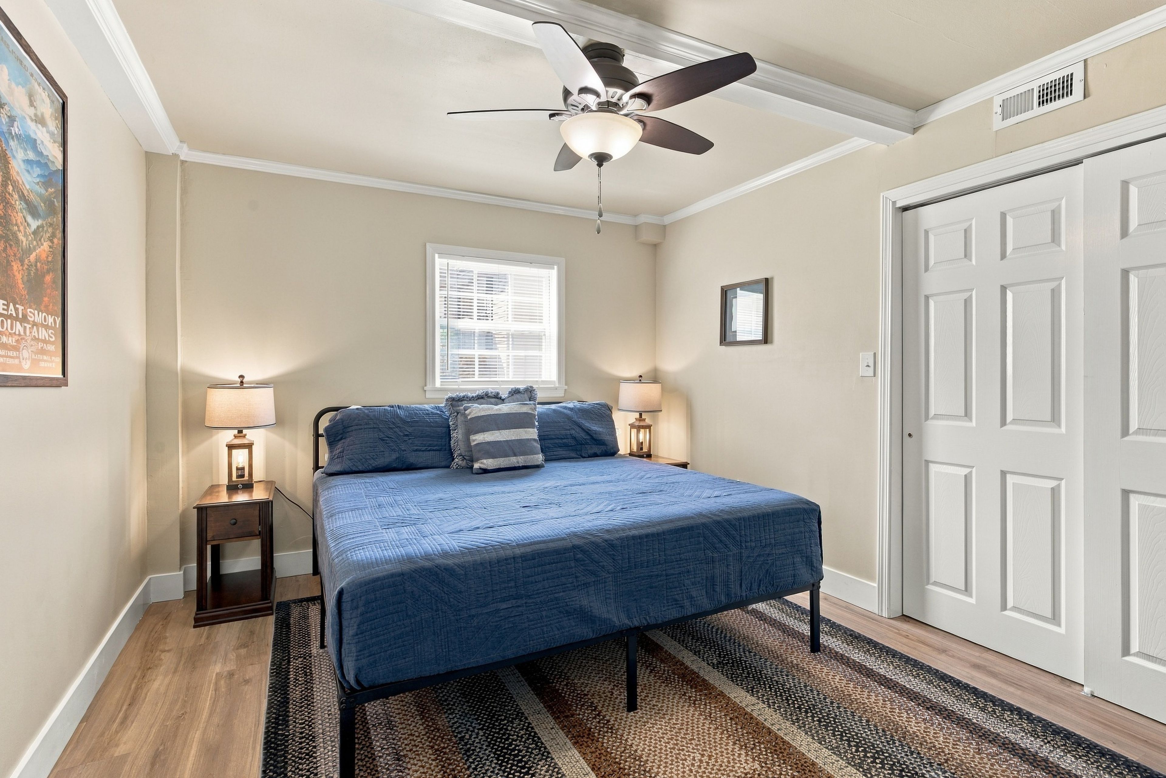 A bedroom with a bed, two bedside lamps, a ceiling fan, a window with blinds, a door, and a poster on the wall.