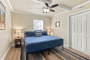 A bedroom with a bed, two bedside lamps, a ceiling fan, a window with blinds, a door, and a poster on the wall.