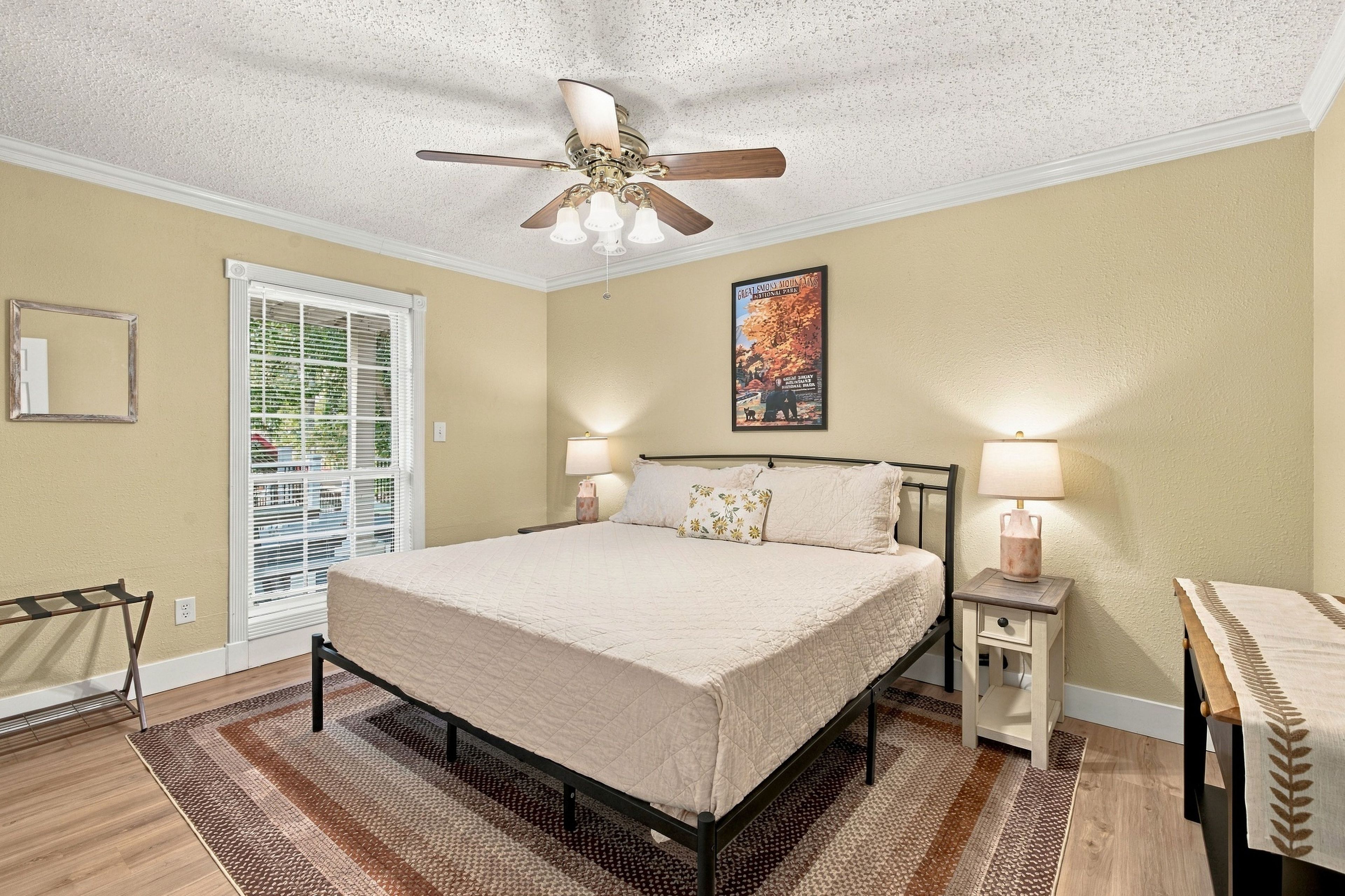A bedroom with a bed, two bedside tables, a ceiling fan, a window with blinds, and a wall-mounted picture.