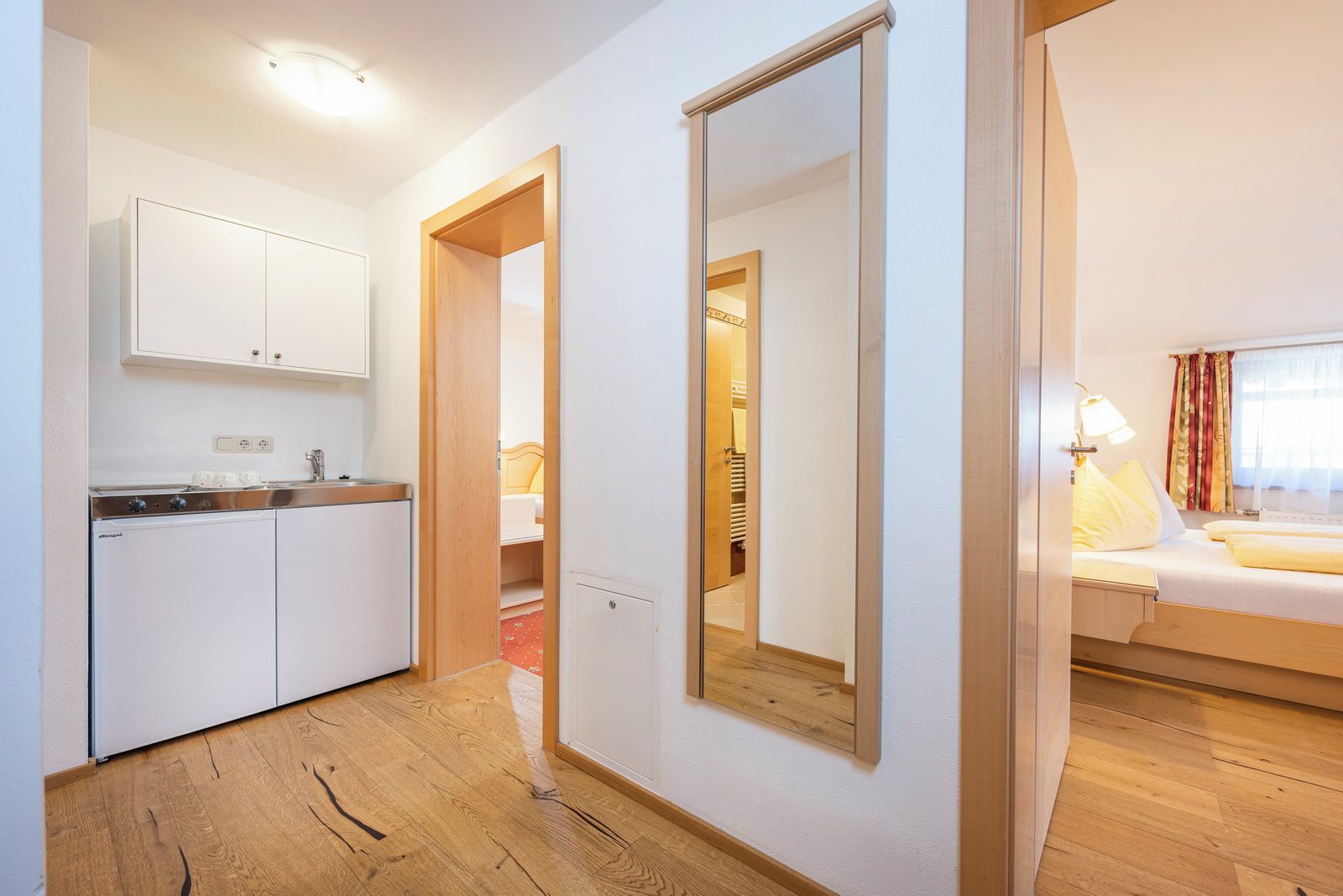 A hotel room with a bed, a bedside lamp, a window with curtains, a wooden floor, a kitchen area with a washing machine, and a mirror.