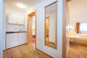 Habitación de hotel con cama, lámpara de noche, ventana con cortinas, suelo de madera, zona de cocina con lavadora y espejo.