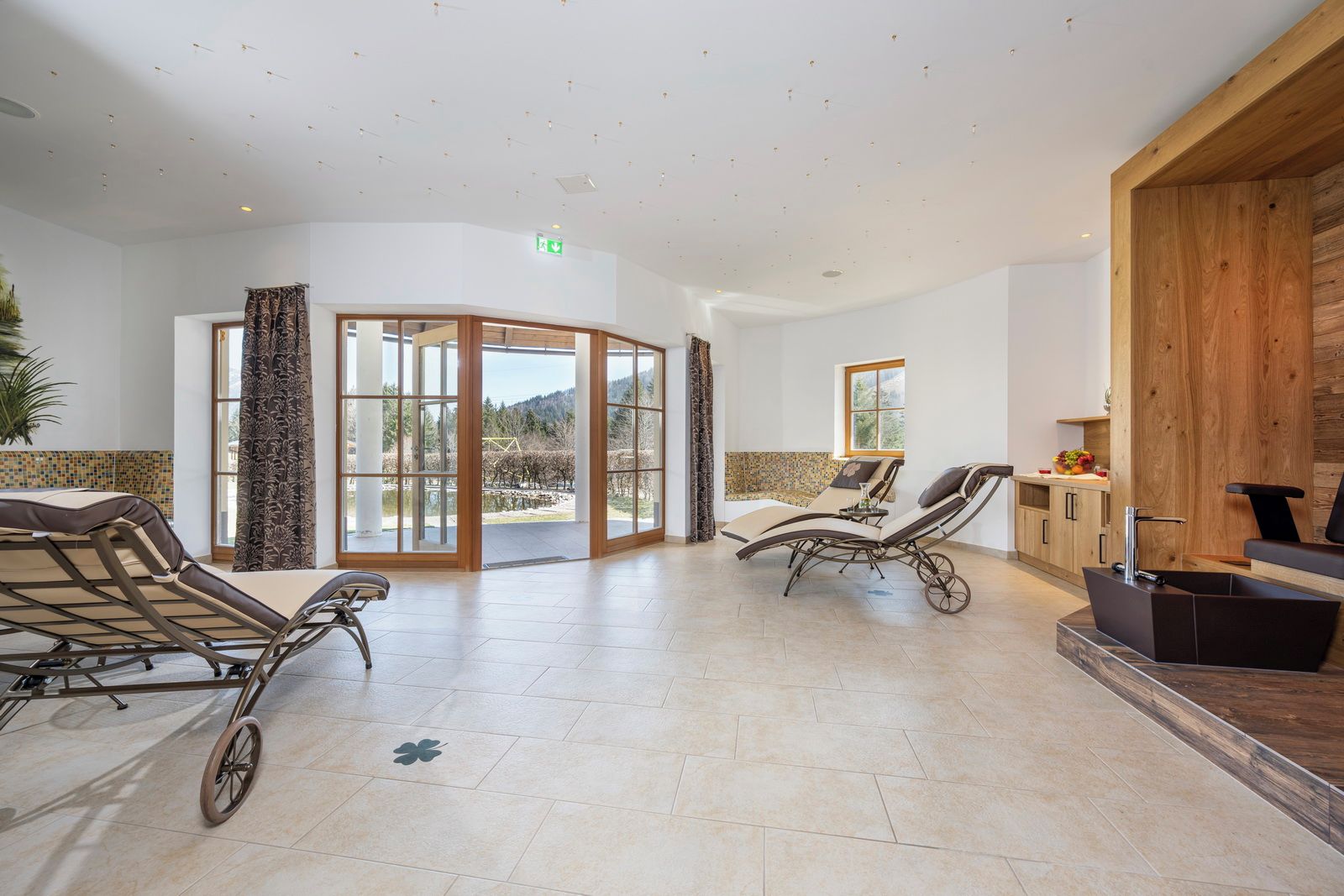 A sunlit room with two reclining chairs, a wooden cabinet, and a view of a vineyard through large glass doors.