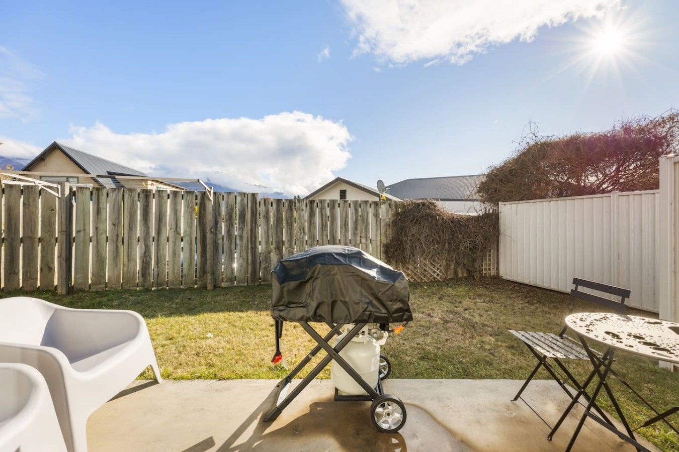 A backyard with a barbecue grill, a white plastic chair, a metal table, and a wooden fence.