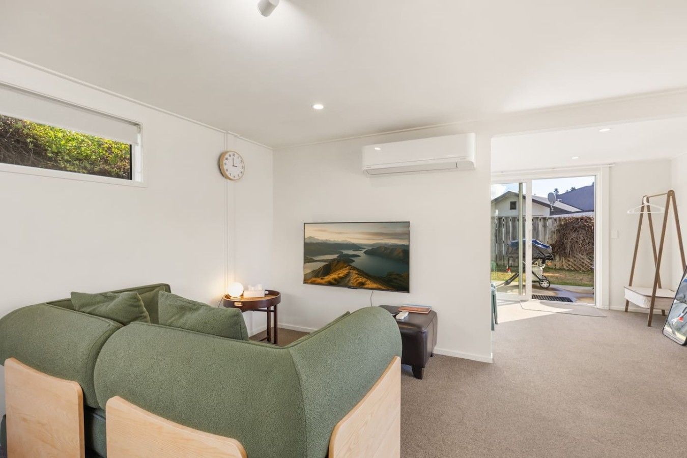 A modern living room with a green sofa, a painting, a clock, and a view of the outdoors through a glass door.