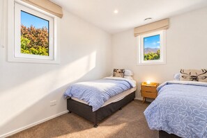 A bedroom with two single beds, a nightstand, and two windows with views of greenery and mountains. - Modern Wānaka Retreat | 4 Bedrooms, Sleeps 8, Hot Tub, 2 Living Areas & Balcony (Wānaka)