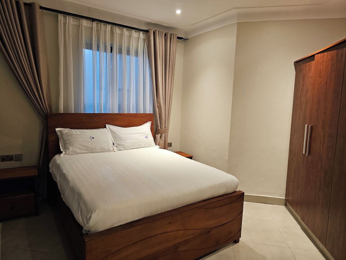 A bedroom with a wooden bed, white bedding, a nightstand, a wardrobe, and sheer curtains.