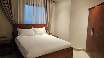 A bedroom with a wooden bed, white bedding, a nightstand, a wardrobe, and sheer curtains.