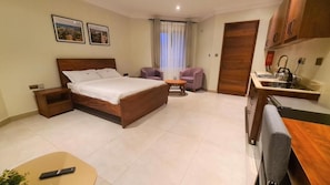 Habitación de hotel con cama, escritorio, silla, mesita, ventana con cortinas y kitchenette.
