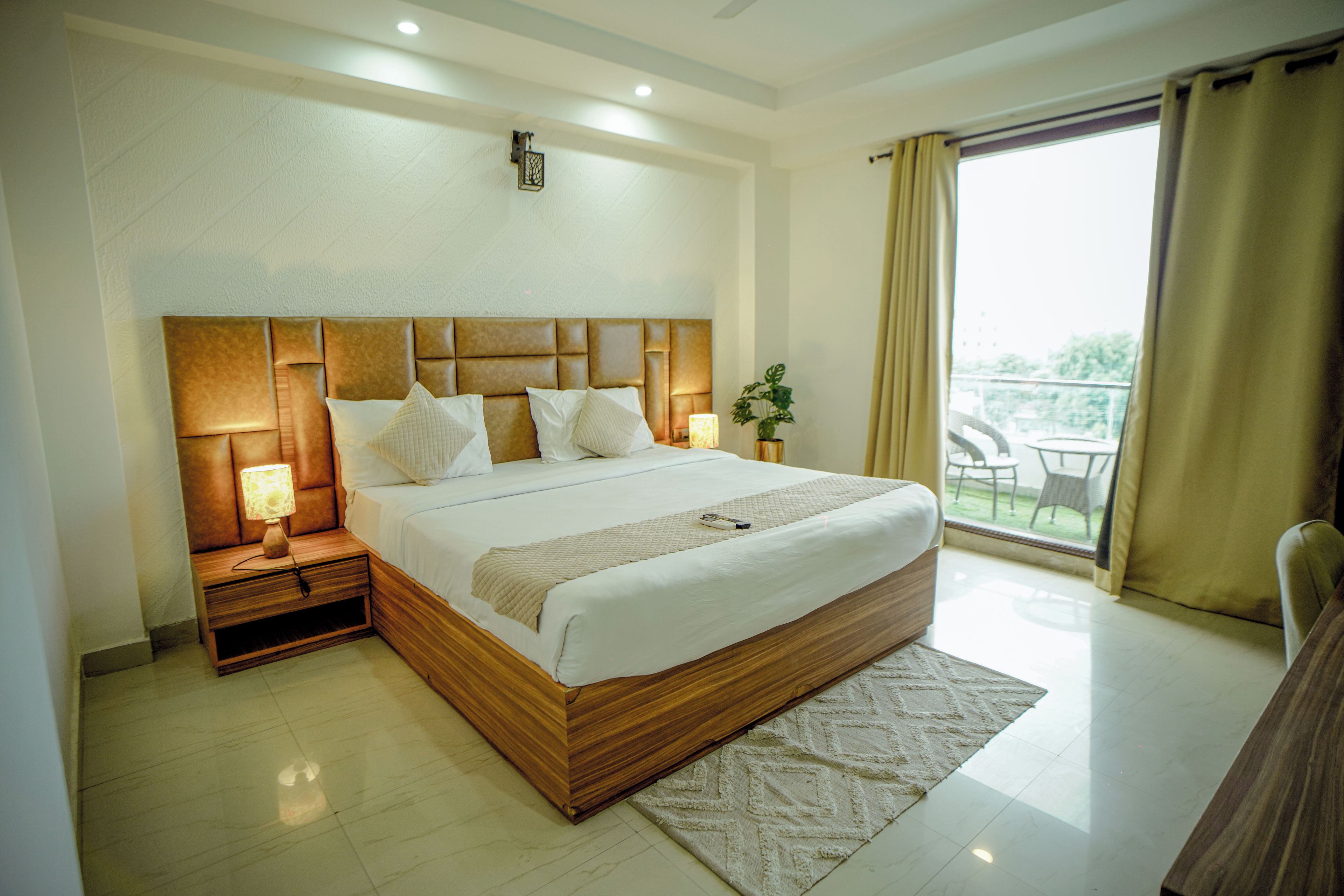 Quarto de hotel com uma cama grande, mesa de cabeceira com luminária, planta em vaso e vista do exterior pela janela.