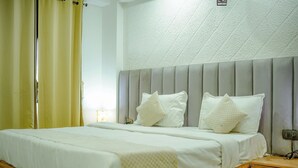 A hotel room with a bed, a bedside table, a nightstand, a wall-mounted light, and a window with curtains. - Hotel Trisha (Gurugram)