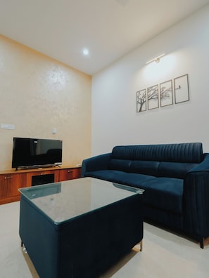 Una sala de estar con un sofá azul oscuro, una mesa de centro de cristal y un televisor de pantalla plana sobre un soporte de madera.