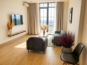 A modern living room with a flat-screen TV, a sofa, a coffee table, and a view through large windows.