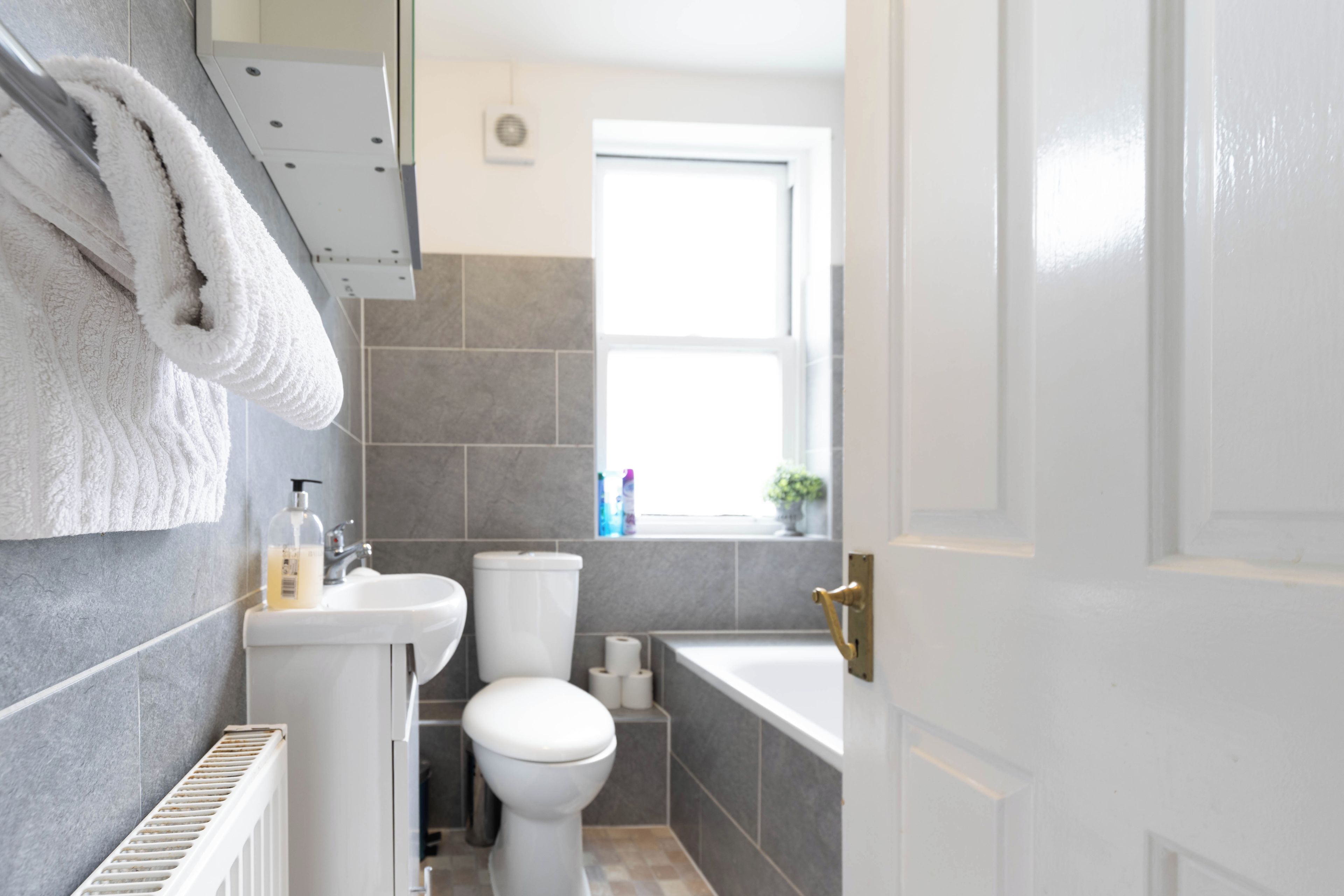 Een badkamer met toilet, wastafel en bad. Er is een handdoekhouder met een handdoek eraan, en een raam boven de wastafel.