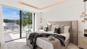 A modern bedroom with a large bed, a glass door leading to a balcony, and a city view. - Vrbo Property (Nueva Andalucía)