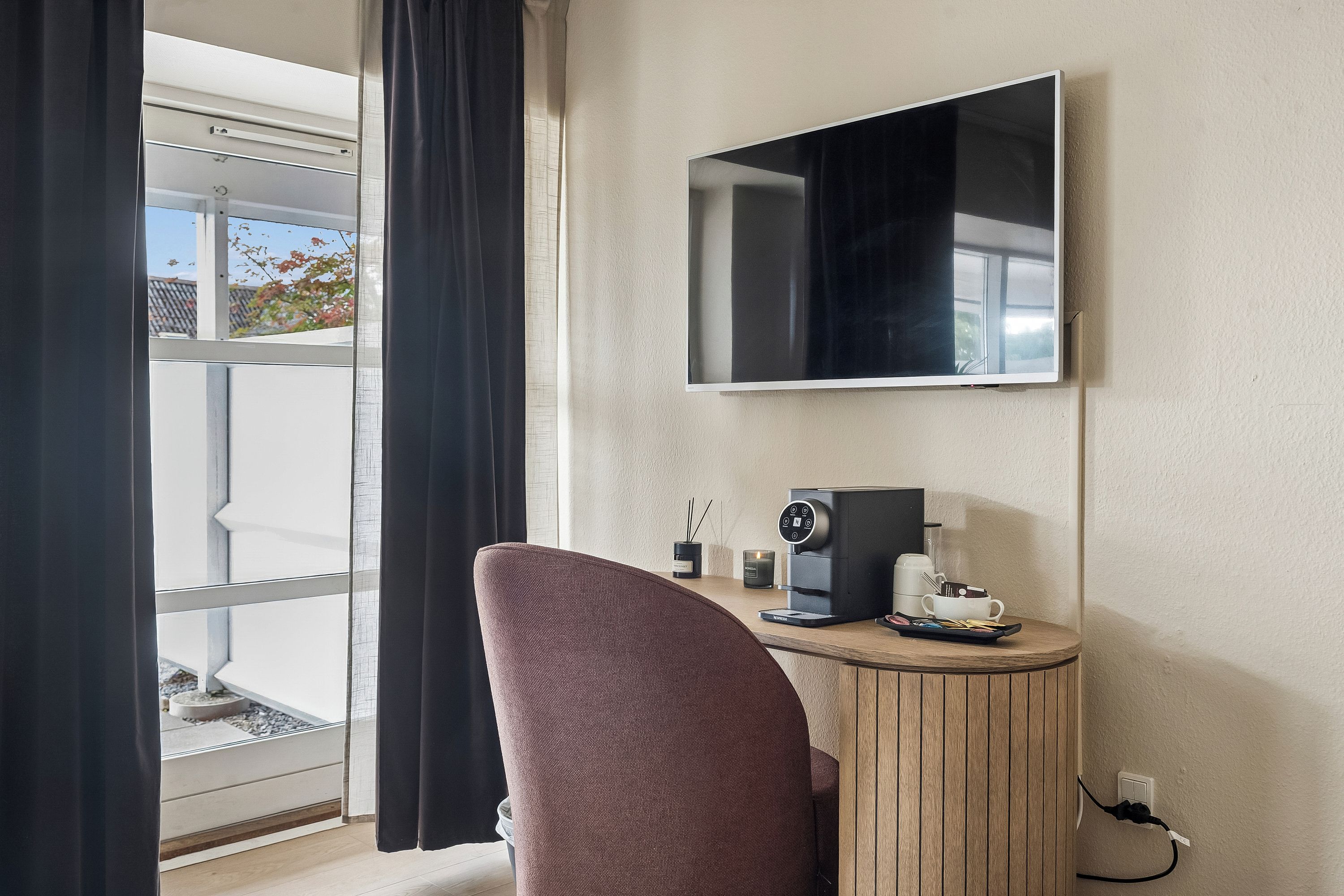 A hotel room with a large window, a flat-screen TV mounted on the wall, a small round table with a coffee machine, and a cushioned chair.