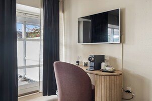 Habitación de hotel con un ventanal grande, un televisor de pantalla plana en la pared, una mesita redonda con una cafetera y un sillón acolchado.