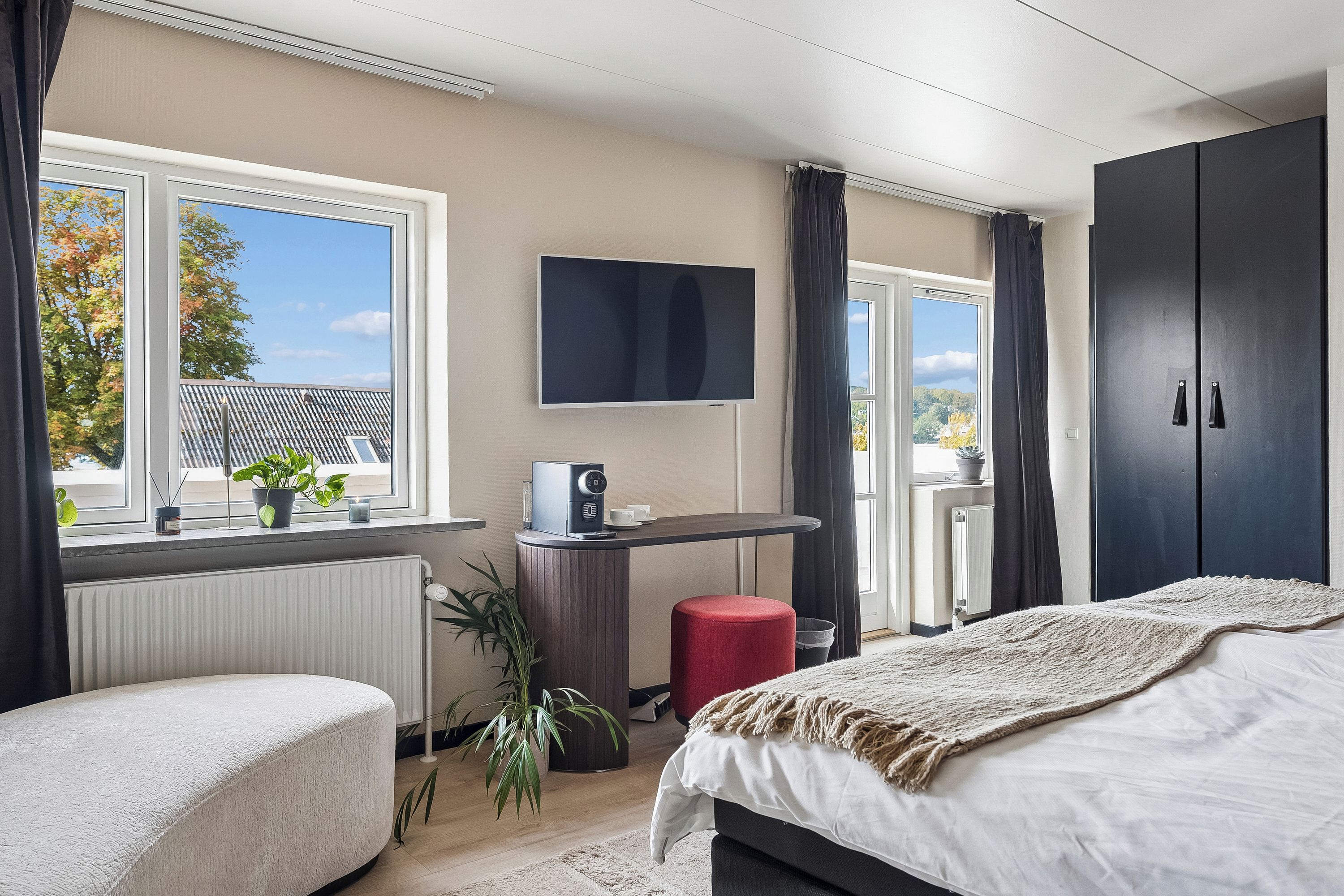 A modern bedroom with a bed, a desk, a TV, and a view of the outdoors.