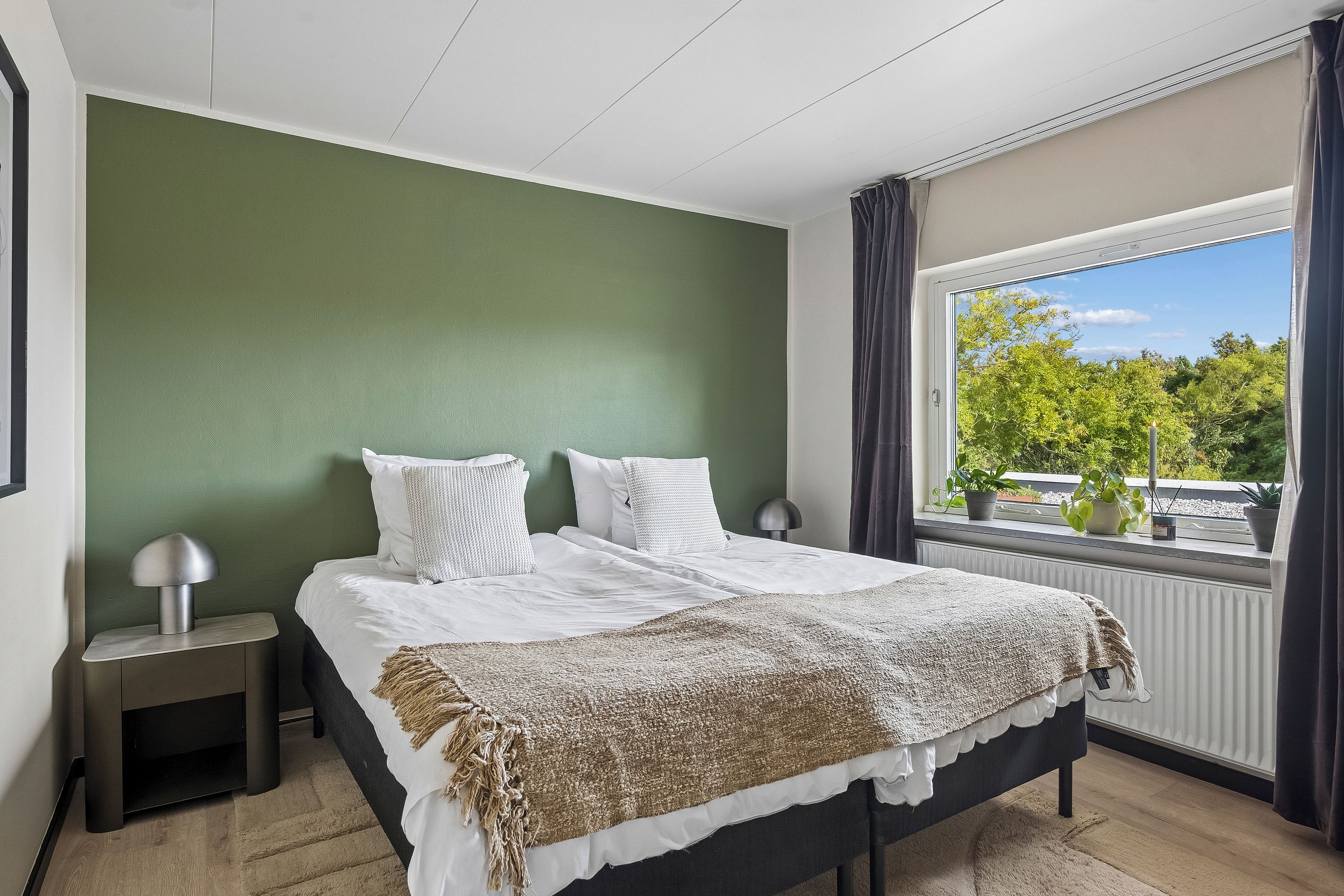 Ein Schlafzimmer mit einem Bett, einem Nachttisch, einer Lampe, einem Fenster mit Vorhängen und Blick auf Grünfläche.