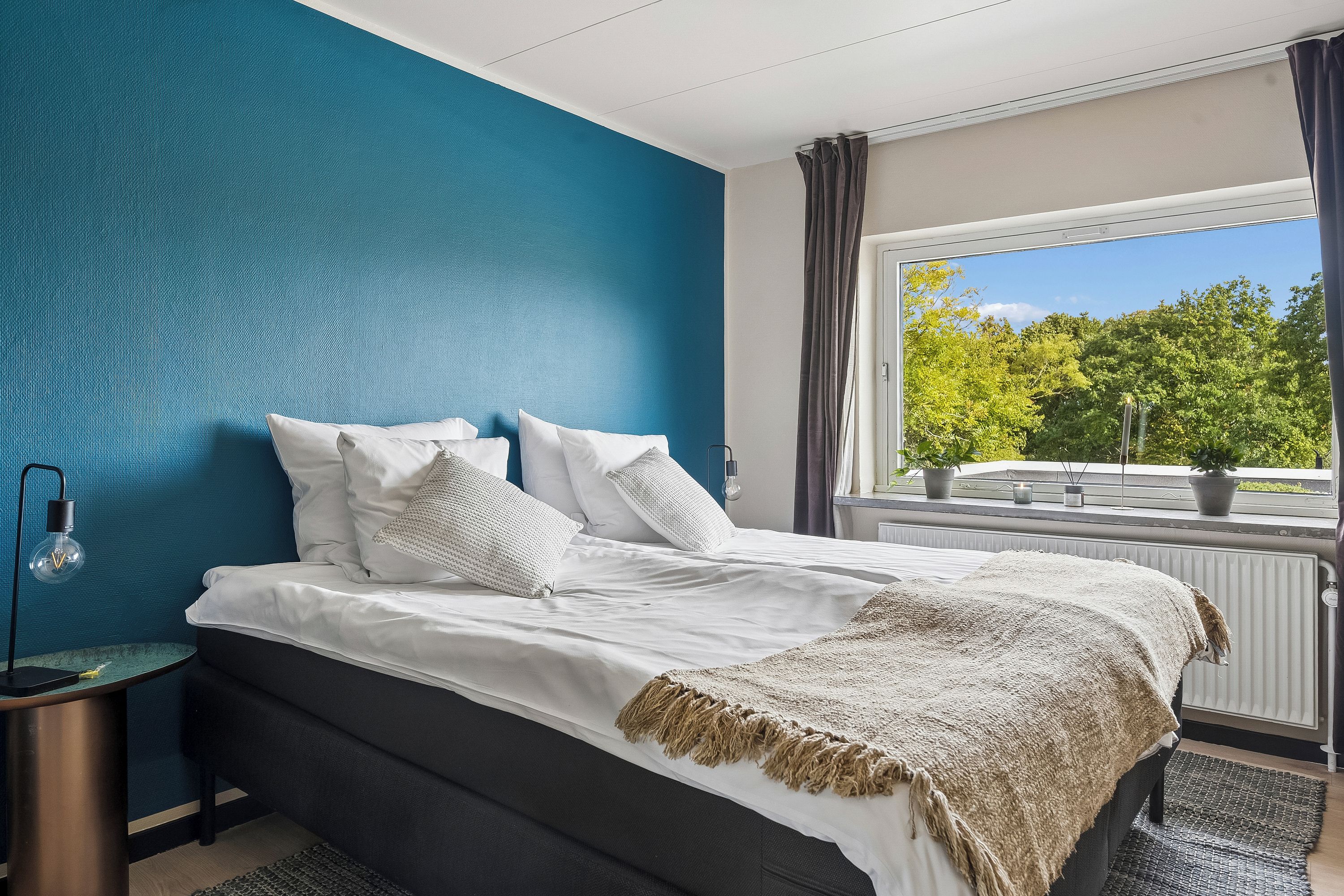 Un dormitorio con cama, mesita de noche, lámpara, ventana con cortinas y vistas a árboles.