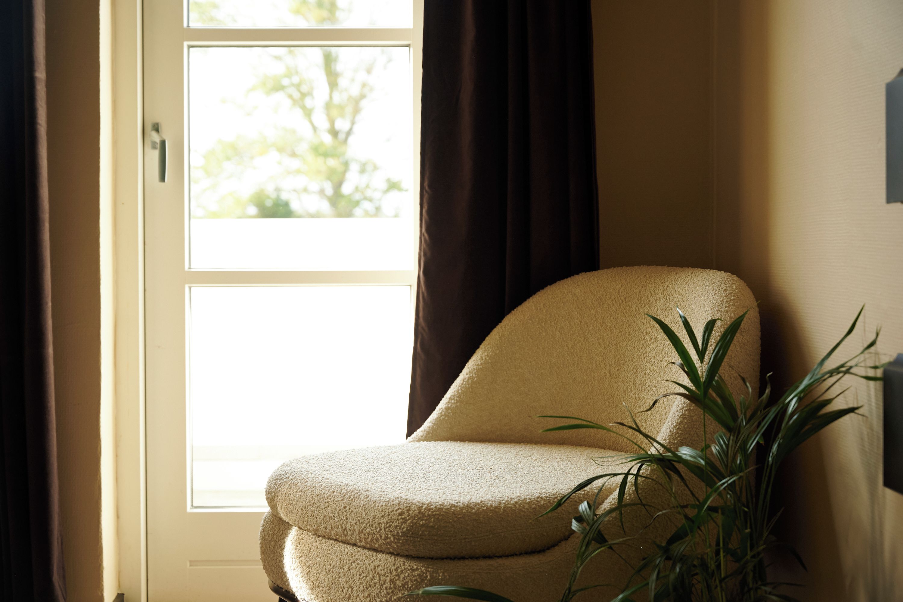 Una silla cerca de una ventana con cortinas, una planta y un aplique en la pared.