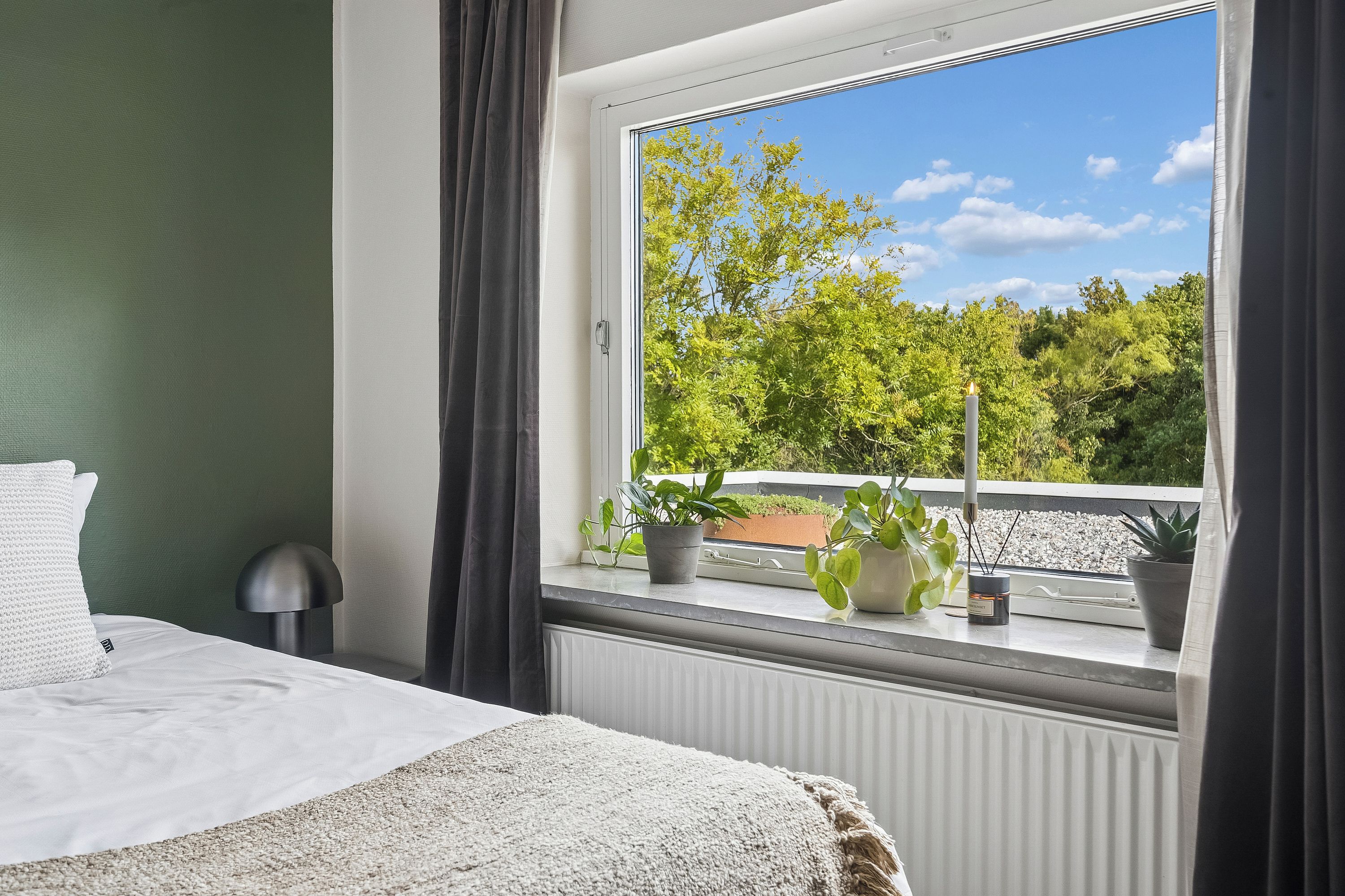 A hotel room with a large window, a bed, a radiator, and several potted plants on the windowsill.