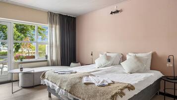 A modern hotel room with a large bed, bedside tables, a window with curtains, and a view of greenery outside.