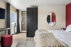 A modern bedroom with a bed, a mirror, a wardrobe, a desk, and a red ottoman.