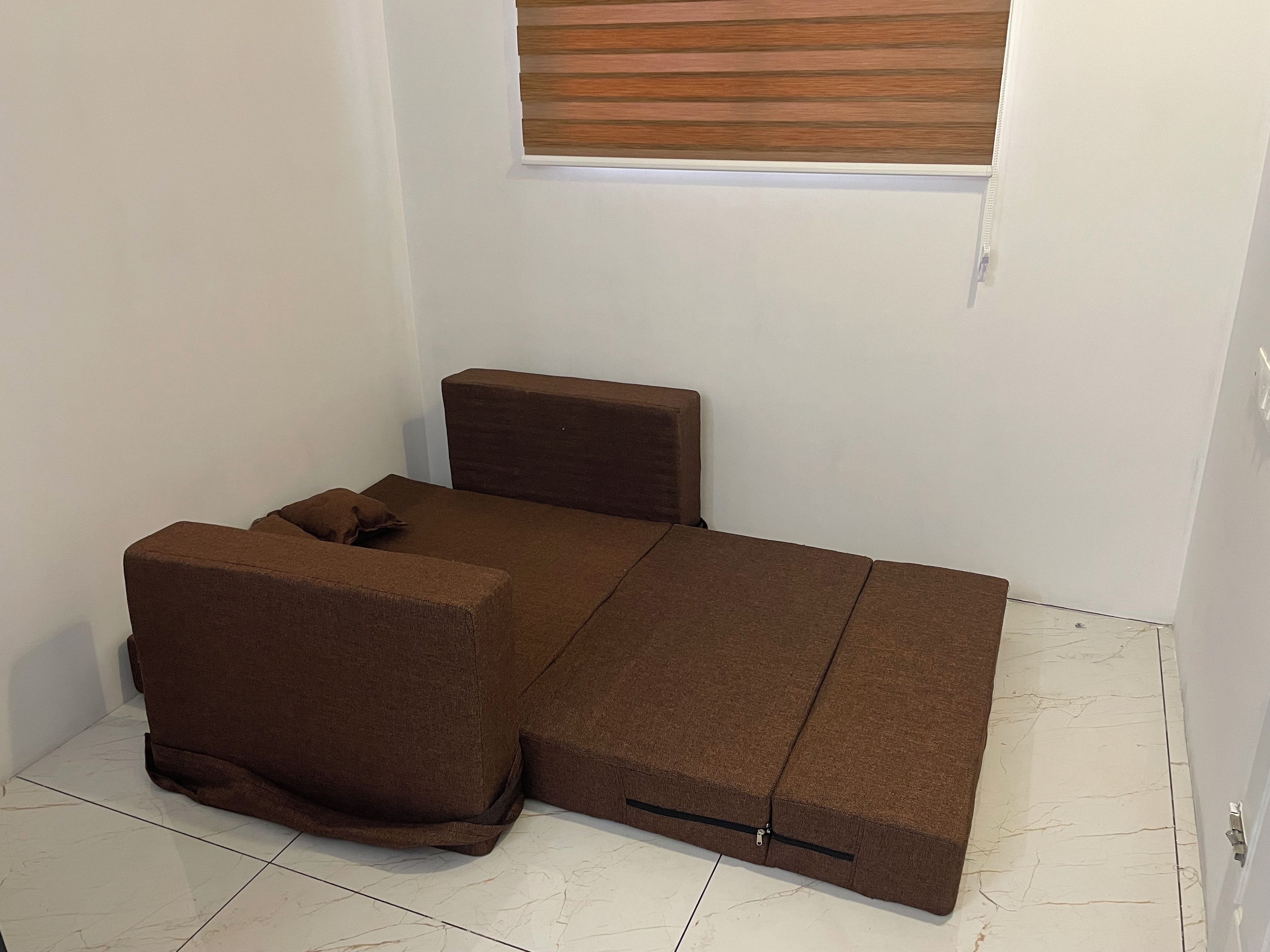 A compact, brown, foldable bed in a room with white walls and a wooden window blind.