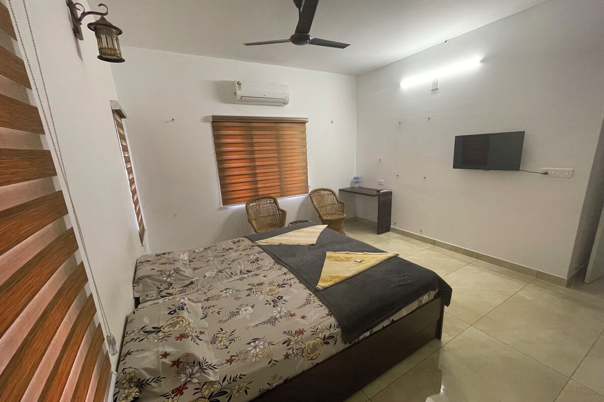 A bedroom with a bed, a desk, a chair, a ceiling fan, and a TV on the wall.