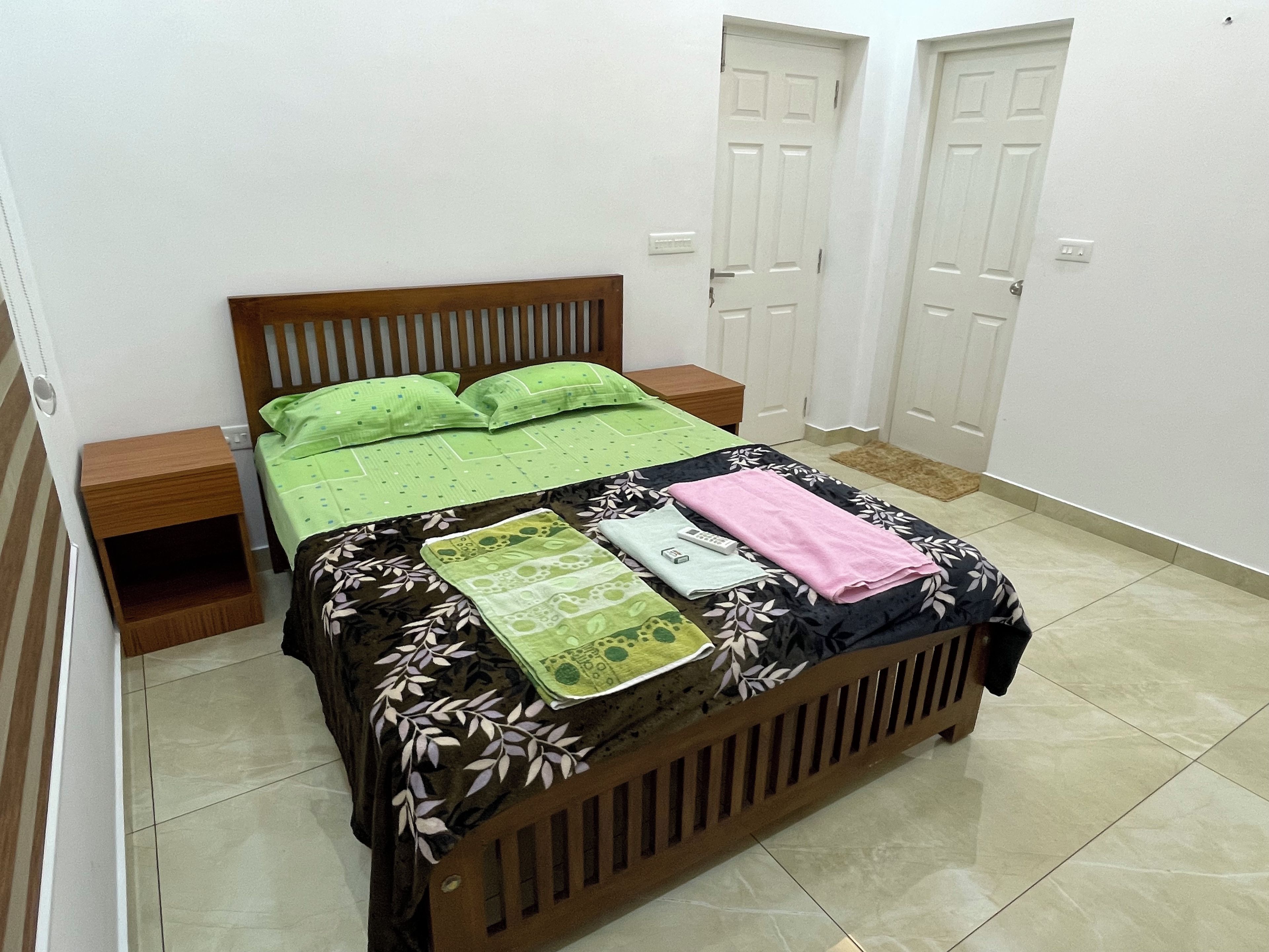 Quarto com cama de madeira, roupa de cama estampada em tons de verde e preto, mesa de cabeceira e criado-mudo.