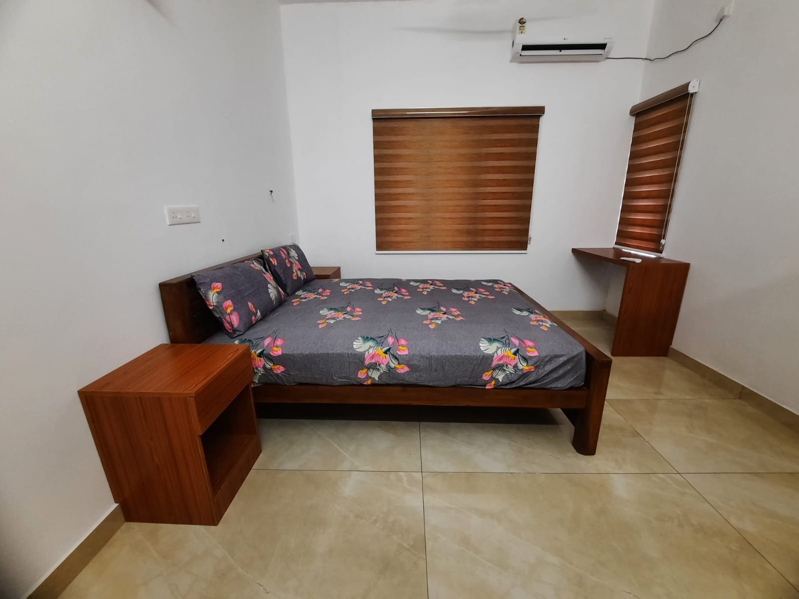 A bedroom with a bed, a nightstand, a wooden wardrobe, a window with blinds, and an air conditioning unit on the wall.