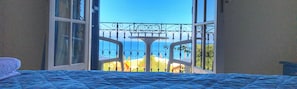 Ein Schlafzimmer mit großem Fenster, einem Bett mit blauer Bettdecke und Blick auf einen Balkon mit Geländer.