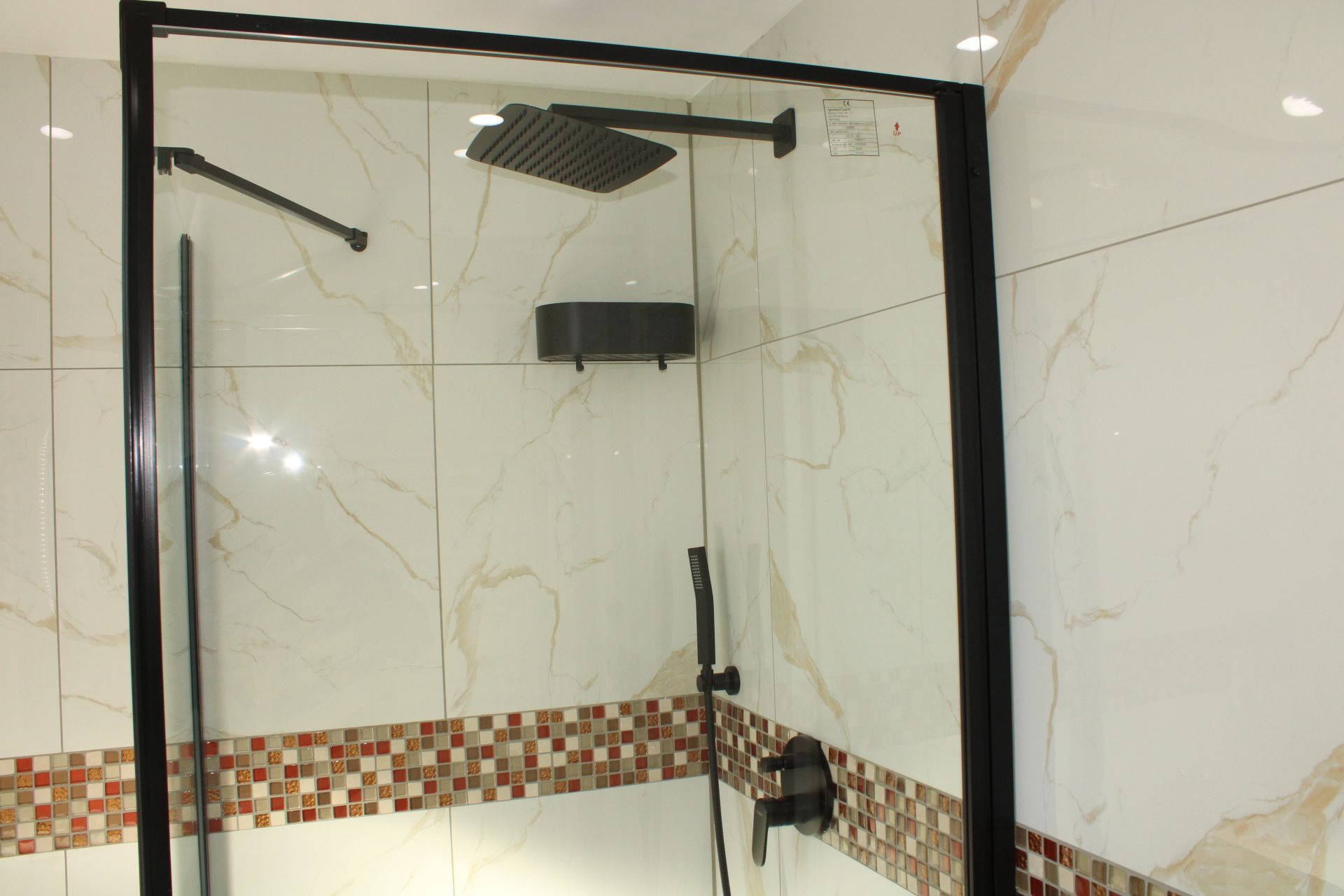Eine gläserne Dusche mit schwarzem Rahmen, einer Handbrause und einer an der Wand befestigten Bedienfeld.