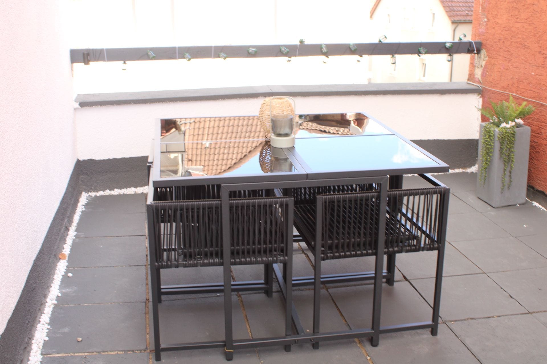 Ein Tisch mit Glasplatte und Metallgestell, begleitet von zwei Stühlen, steht auf einer Terrasse mit gefliestem Boden.